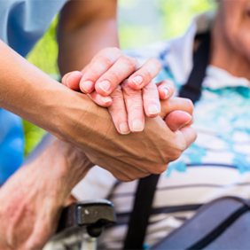 Auf dem Bild sieht man eine Nahaufnahme von zwei Händen, die sich halten. Eine Hand gehört zu einer älteren Person, die in einem Rollstuhl sitzt und ein gestreiftes Hemd trägt. Die andere Hand gehört zu einer jüngeren Person, die Pflege leistet. Im Hintergrund ist das Gesicht der älteren Person leicht unscharf zu erkennen, sie scheint zu lächeln.
