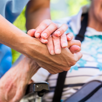 Auf dem Bild sieht man eine Nahaufnahme von zwei Händen, die sich halten. Eine Hand gehört zu einer älteren Person, die in einem Rollstuhl sitzt und ein gestreiftes Hemd trägt. Die andere Hand gehört zu einer jüngeren Person, die Pflege leistet. Im Hintergrund ist das Gesicht der älteren Person leicht unscharf zu erkennen, sie scheint zu lächeln.