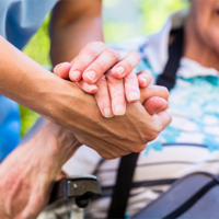 Auf dem Bild sieht man eine Nahaufnahme von zwei Händen, die sich halten. Eine Hand gehört zu einer älteren Person, die in einem Rollstuhl sitzt und ein gestreiftes Hemd trägt. Die andere Hand gehört zu einer jüngeren Person, die Pflege leistet. Im Hintergrund ist das Gesicht der älteren Person leicht unscharf zu erkennen, sie scheint zu lächeln.