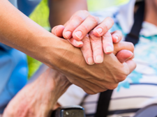 Auf dem Bild sieht man eine Nahaufnahme von zwei Händen, die sich halten. Eine Hand gehört zu einer älteren Person, die in einem Rollstuhl sitzt und ein gestreiftes Hemd trägt. Die andere Hand gehört zu einer jüngeren Person, die Pflege leistet. Im Hintergrund ist das Gesicht der älteren Person leicht unscharf zu erkennen, sie scheint zu lächeln.