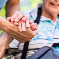 Auf dem Bild sieht man eine Nahaufnahme von zwei Händen, die sich halten. Eine Hand gehört zu einer älteren Person, die in einem Rollstuhl sitzt und ein gestreiftes Hemd trägt. Die andere Hand gehört zu einer jüngeren Person, die Pflege leistet. Im Hintergrund ist das Gesicht der älteren Person leicht unscharf zu erkennen, sie scheint zu lächeln.