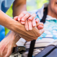 Auf dem Bild sieht man eine Nahaufnahme von zwei Händen, die sich halten. Eine Hand gehört zu einer älteren Person, die in einem Rollstuhl sitzt und ein gestreiftes Hemd trägt. Die andere Hand gehört zu einer jüngeren Person, die Pflege leistet. Im Hintergrund ist das Gesicht der älteren Person leicht unscharf zu erkennen, sie scheint zu lächeln.