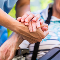Auf dem Bild sieht man eine Nahaufnahme von zwei Händen, die sich halten. Eine Hand gehört zu einer älteren Person, die in einem Rollstuhl sitzt und ein gestreiftes Hemd trägt. Die andere Hand gehört zu einer jüngeren Person, die Pflege leistet. Im Hintergrund ist das Gesicht der älteren Person leicht unscharf zu erkennen, sie scheint zu lächeln.