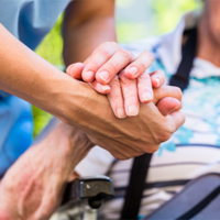 Auf dem Bild sieht man eine Nahaufnahme von zwei Händen, die sich halten. Eine Hand gehört zu einer älteren Person, die in einem Rollstuhl sitzt und ein gestreiftes Hemd trägt. Die andere Hand gehört zu einer jüngeren Person, die Pflege leistet. Im Hintergrund ist das Gesicht der älteren Person leicht unscharf zu erkennen, sie scheint zu lächeln.