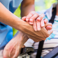 Auf dem Bild sieht man eine Nahaufnahme von zwei Händen, die sich halten. Eine Hand gehört zu einer älteren Person, die in einem Rollstuhl sitzt und ein gestreiftes Hemd trägt. Die andere Hand gehört zu einer jüngeren Person, die Pflege leistet. Im Hintergrund ist das Gesicht der älteren Person leicht unscharf zu erkennen, sie scheint zu lächeln.