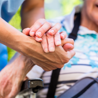 Auf dem Bild sieht man eine Nahaufnahme von zwei Händen, die sich halten. Eine Hand gehört zu einer älteren Person, die in einem Rollstuhl sitzt und ein gestreiftes Hemd trägt. Die andere Hand gehört zu einer jüngeren Person, die Pflege leistet. Im Hintergrund ist das Gesicht der älteren Person leicht unscharf zu erkennen, sie scheint zu lächeln.