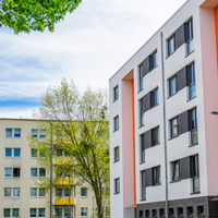 Das Bild zeigt eine städtische Wohngegend mit modernen Mehrfamilienhäusern. Die Gebäude haben eine helle Fassade, wobei eines der Häuser durch orangefarbene Akzente hervorsticht. Die Fenster sind gleichmäßig angeordnet und einige Balkone sind zu sehen. 