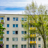 Das Bild zeigt eine städtische Wohngegend mit modernen Mehrfamilienhäusern. Die Gebäude haben eine helle Fassade, wobei eines der Häuser durch orangefarbene Akzente hervorsticht. Die Fenster sind gleichmäßig angeordnet und einige Balkone sind zu sehen. 
