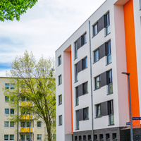 Das Bild zeigt eine städtische Wohngegend mit modernen Mehrfamilienhäusern. Die Gebäude haben eine helle Fassade, wobei eines der Häuser durch orangefarbene Akzente hervorsticht. Die Fenster sind gleichmäßig angeordnet und einige Balkone sind zu sehen. 