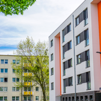 Das Bild zeigt eine städtische Wohngegend mit modernen Mehrfamilienhäusern. Die Gebäude haben eine helle Fassade, wobei eines der Häuser durch orangefarbene Akzente hervorsticht. Die Fenster sind gleichmäßig angeordnet und einige Balkone sind zu sehen. 