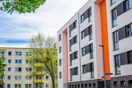 Das Bild zeigt eine städtische Wohngegend mit modernen Mehrfamilienhäusern. Die Gebäude haben eine helle Fassade, wobei eines der Häuser durch orangefarbene Akzente hervorsticht. Die Fenster sind gleichmäßig angeordnet und einige Balkone sind zu sehen. 