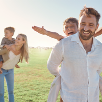 Auf dem Bild ist eine Familie zu sehen, die auf einer Wiese sind. Im Hintergrund trägt die Frau ein Kind auf dem Rücken. Der Mann im Vordergrund trägt auch ein Kind auf dem Rücken. Das Kind im Vordergrund streckt die Arme aus. 
