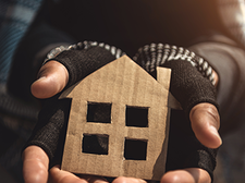 Eine Person hält ein kleines, aus Pappe ausgeschnittenes Symbol in Form eines Hauses in ihren Händen. Die Hände tragen fingerlose Handschuhe, die auf Kälte und möglicherweise Wohnungslosigkeit hinweisen. Die Kleidung wirkt abgenutzt. Das Licht fällt warm von der Seite ein und beleuchtet das Haus-Symbol.