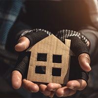 Eine Person hält ein kleines, aus Pappe ausgeschnittenes Symbol in Form eines Hauses in ihren Händen. Die Hände tragen fingerlose Handschuhe, die auf Kälte und möglicherweise Wohnungslosigkeit hinweisen. Die Kleidung wirkt abgenutzt. Das Licht fällt warm von der Seite ein und beleuchtet das Haus-Symbol.