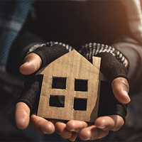Eine Person hält ein kleines, aus Pappe ausgeschnittenes Symbol in Form eines Hauses in ihren Händen. Die Hände tragen fingerlose Handschuhe, die auf Kälte und möglicherweise Wohnungslosigkeit hinweisen. Die Kleidung wirkt abgenutzt. Das Licht fällt warm von der Seite ein und beleuchtet das Haus-Symbol.
