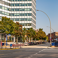 Eine breite, mehrspurige Straße verläuft durch ein urbanes Gebiet mit modernen Bürogebäuden und Bäumen am Straßenrand. Links ist ein Eingang zur U-Bahn-Station erkennbar. Mehrere geparkte Autos stehen am Straßenrand, daneben befinden sich Fahrradständer mit Leihfahrrädern. Rechts ragt ein rotes Backsteingebäude mit Glasfassade in das Bild. Die Straße ist fast leer, der Himmel ist blau und klar. 
