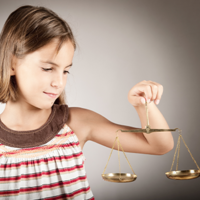 Ein junges Mädchen mit schulterlangem braunem Haar hält eine goldene Waage in ihrer rechten Hand. Sie trägt ein ärmelloses Oberteil mit Streifen in verschiedenen Farben und schaut konzentriert auf die Waage. Der Hintergrund ist neutral und grau.