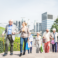 Eine Gruppe älterer Menschen geht eine Treppe hinauf. Sie tragen bunte Einkaufstaschen. Im Hintergrund sind moderne Bürogebäude und grüne Pflanzen zu sehen. Die Sonne scheint. Die Personen unterstützen sich gegenseitig beim Gehen.