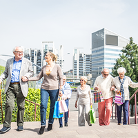 Eine Gruppe älterer Menschen geht eine Treppe hinauf. Sie tragen bunte Einkaufstaschen. Im Hintergrund sind moderne Bürogebäude und grüne Pflanzen zu sehen. Die Sonne scheint. Die Personen unterstützen sich gegenseitig beim Gehen.