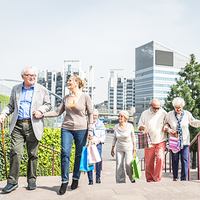 Eine Gruppe älterer Menschen geht eine Treppe hinauf. Sie tragen bunte Einkaufstaschen. Im Hintergrund sind moderne Bürogebäude und grüne Pflanzen zu sehen. Die Sonne scheint. Die Personen unterstützen sich gegenseitig beim Gehen.