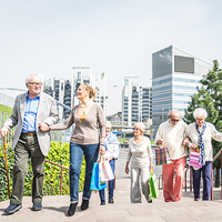 Eine Gruppe älterer Menschen geht eine Treppe hinauf. Sie tragen bunte Einkaufstaschen. Im Hintergrund sind moderne Bürogebäude und grüne Pflanzen zu sehen. Die Sonne scheint. Die Personen unterstützen sich gegenseitig beim Gehen.