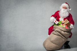 Ein Mann in einem traditionellen Weihnachtsmannkostüm steht vor einem grauen Hintergrund. Neben ihm steht ein großer, brauner Sack, der mit verschiedenen Geschenken gefüllt ist. Man kann bunte Geschenkverpackungen und ein Stofftier erkennen, die aus dem Sack herausragen.