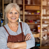 Ältere Frau mit grauen Haaren steht lächelnd in einer Werkstatt oder einem Atelier. Sie trägt ein graues Oberteil und eine braune Schürze und hat die Arme vor der Brust verschränkt. Im Hintergrund sind Regale mit Materialien und Werkzeugen zu sehen.