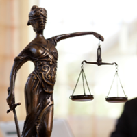 Das Bild zeigt eine Frau in einem Büro, die an einem Laptop arbeitet. Im Vordergrund steht eine Statue der Justitia, die Göttin der Gerechtigkeit, mit einer Waage in der einen und einem Schwert in der anderen Hand. Die Frau im Hintergrund ist unscharf dargestellt, während die Statue scharf fokussiert ist. Auf dem Schreibtisch vor ihr liegt ein aufgeschlagenes Buch. 