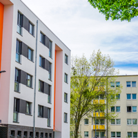 Das Bild zeigt eine städtische Wohngegend mit modernen Mehrfamilienhäusern. Die Gebäude haben eine helle Fassade, wobei eines der Häuser durch orangefarbene Akzente hervorsticht. Die Fenster sind gleichmäßig angeordnet und einige Balkone sind zu sehen. 