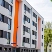 Das Bild zeigt eine städtische Wohngegend mit modernen Mehrfamilienhäusern. Die Gebäude haben eine helle Fassade, wobei eines der Häuser durch orangefarbene Akzente hervorsticht. Die Fenster sind gleichmäßig angeordnet und einige Balkone sind zu sehen. 