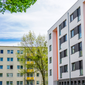Das Bild zeigt eine städtische Wohngegend mit modernen Mehrfamilienhäusern. Die Gebäude haben eine helle Fassade, wobei eines der Häuser durch orangefarbene Akzente hervorsticht. Die Fenster sind gleichmäßig angeordnet und einige Balkone sind zu sehen. 