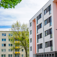 Das Bild zeigt eine städtische Wohngegend mit modernen Mehrfamilienhäusern. Die Gebäude haben eine helle Fassade, wobei eines der Häuser durch orangefarbene Akzente hervorsticht. Die Fenster sind gleichmäßig angeordnet und einige Balkone sind zu sehen. 