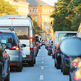 Das Bild zeigt eine Straße, die von dichtem Verkehr geprägt ist. Autos und Lieferwagen stehen eng zusammen in beiden Fahrtrichtungen. Die Fahrzeuge sind unterschiedlich groß und in verschiedenen Farben lackiert. Im Hintergrund sind mehrstöckige Gebäude zu sehen, die von Bäumen gesäumt werden. Die Sonne scheint. Ein grünes Ampellicht ist im Hintergrund sichtbar.