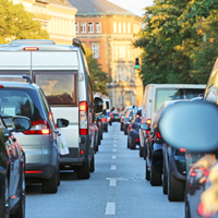 Das Bild zeigt eine Straße, die von dichtem Verkehr geprägt ist. Autos und Lieferwagen stehen eng zusammen in beiden Fahrtrichtungen. Die Fahrzeuge sind unterschiedlich groß und in verschiedenen Farben lackiert. Im Hintergrund sind mehrstöckige Gebäude zu sehen, die von Bäumen gesäumt werden. Die Sonne scheint. Ein grünes Ampellicht ist im Hintergrund sichtbar.