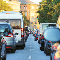 Das Bild zeigt eine Straße, die von dichtem Verkehr geprägt ist. Autos und Lieferwagen stehen eng zusammen in beiden Fahrtrichtungen. Die Fahrzeuge sind unterschiedlich groß und in verschiedenen Farben lackiert. Im Hintergrund sind mehrstöckige Gebäude zu sehen, die von Bäumen gesäumt werden. Die Sonne scheint. Ein grünes Ampellicht ist im Hintergrund sichtbar.