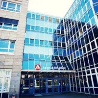 Das Bild zeigt die Fassade eines modernen Bürogebäudes mit großen Glasfenstern. Das Gebäude hat mehrere Stockwerke und eine Kombination aus blauen Glasflächen und beigen Steinwänden. Über dem Eingang befindet sich ein Schild mit der Aufschrift "Agentur für Arbeit" und einem roten Logo. Der Eingangsbereich ist durch eine Glasüberdachung geschützt, und es gibt mehrere Türen, die in das Gebäude führen. 