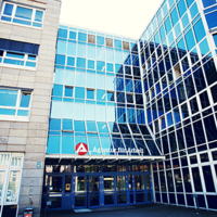Das Bild zeigt die Fassade eines modernen Bürogebäudes mit großen Glasfenstern. Das Gebäude hat mehrere Stockwerke und eine Kombination aus blauen Glasflächen und beigen Steinwänden. Über dem Eingang befindet sich ein Schild mit der Aufschrift "Agentur für Arbeit" und einem roten Logo. Der Eingangsbereich ist durch eine Glasüberdachung geschützt, und es gibt mehrere Türen, die in das Gebäude führen. 