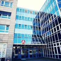 Das Bild zeigt die Fassade eines modernen Bürogebäudes mit großen Glasfenstern. Das Gebäude hat mehrere Stockwerke und eine Kombination aus blauen Glasflächen und beigen Steinwänden. Über dem Eingang befindet sich ein Schild mit der Aufschrift "Agentur für Arbeit" und einem roten Logo. Der Eingangsbereich ist durch eine Glasüberdachung geschützt, und es gibt mehrere Türen, die in das Gebäude führen. 