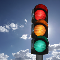Das Bild zeigt eine Ampel vor einem Hintergrund mit blauem Himmel und einigen weißen Wolken. Die Ampel hat drei Lichter: das obere Licht leuchtet rot, das mittlere Licht leuchtet gelb, und das untere Licht leuchtet grün.