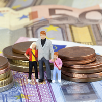 Das Bild zeigt drei Miniaturfiguren, die eine Familie darstellen, bestehend aus einem Erwachsenen und zwei Kindern. Sie stehen auf einem Haufen von Euro-Münzen und -Scheinen. Die Münzen sind in verschiedenen Größen gestapelt, und die Scheine sind im Hintergrund sichtbar. 