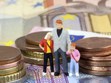 Das Bild zeigt drei Miniaturfiguren, die eine Familie darstellen, bestehend aus einem Erwachsenen und zwei Kindern. Sie stehen auf einem Haufen von Euro-Münzen und -Scheinen. Die Münzen sind in verschiedenen Größen gestapelt, und die Scheine sind im Hintergrund sichtbar.