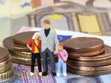 Das Bild zeigt drei Miniaturfiguren, die eine Familie darstellen, bestehend aus einem Erwachsenen und zwei Kindern. Sie stehen auf einem Haufen von Euro-Münzen und -Scheinen. Die Münzen sind in verschiedenen Größen gestapelt, und die Scheine sind im Hintergrund sichtbar.