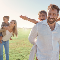 Auf dem Bild ist eine Familie zu sehen, die auf einer Wiese sind. Im Hintergrund trägt die Frau ein Kind auf dem Rücken. Der Mann im Vordergrund trägt auch ein Kind auf dem Rücken. Das Kind im Vordergrund streckt die Arme aus. 