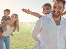 Auf dem Bild ist eine Familie zu sehen, die auf einer Wiese sind. Im Hintergrund trägt die Frau ein Kind auf dem Rücken. Der Mann im Vordergrund trägt auch ein Kind auf dem Rücken. Das Kind im Vordergrund streckt die Arme aus.