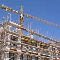Das Bild zeigt eine Baustelle, auf der ein mehrstöckiges Gebäude errichtet wird. Das Gebäude ist von einem Gerüst umgeben, das den Arbeitern Zugang zu verschiedenen Ebenen ermöglicht. Im Hintergrund ragt ein großer Baukran in den klaren blauen Himmel, der für den Transport von Baumaterialien verwendet wird. 