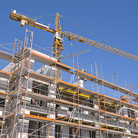 Das Bild zeigt eine Baustelle, auf der ein mehrstöckiges Gebäude errichtet wird. Das Gebäude ist von einem Gerüst umgeben, das den Arbeitern Zugang zu verschiedenen Ebenen ermöglicht. Im Hintergrund ragt ein großer Baukran in den klaren blauen Himmel, der für den Transport von Baumaterialien verwendet wird. 