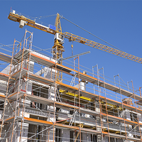 Das Bild zeigt eine Baustelle, auf der ein mehrstöckiges Gebäude errichtet wird. Das Gebäude ist von einem Gerüst umgeben, das den Arbeitern Zugang zu verschiedenen Ebenen ermöglicht. Im Hintergrund ragt ein großer Baukran in den klaren blauen Himmel, der für den Transport von Baumaterialien verwendet wird. 