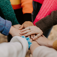Eine Gruppe von Menschen steht im Kreis und legt ihre Hände übereinander in die Mitte. Sie tragen bunte Pullover in verschiedenen Farben wie Pink, Orange, Grün und Blau. 