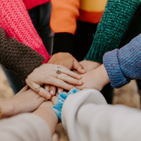 Eine Gruppe von Menschen steht im Kreis und legt ihre Hände übereinander in die Mitte. Sie tragen bunte Pullover in verschiedenen Farben wie Pink, Orange, Grün und Blau. 