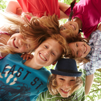 Auf dem Bild sind sechs fröhliche Kinder zu sehen, die in einem Kreis stehen und ihre Köpfe zusammenstecken. Sie lachen und schauen nach unten in die Kamera. Die Kinder tragen bunte Kleidung. Im Hintergrund sind grüne Blätter von Bäumen zu erkennen. 